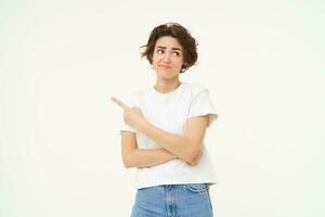 Image of woman with sceptical expression, pointing finger left, showing advertisement with doubtful face expression, standing over white studio background photo