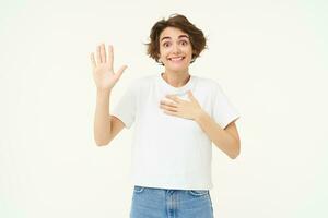 Portrait of beautiful young woman makes promise, raises one hand and puts one on heart, tells truth, gives oath, stands over white background photo