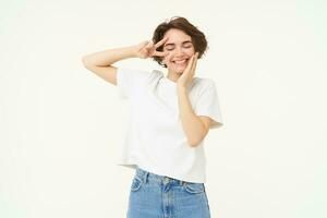 Portrait of young girl shows peace, v-sign and smiling, express posivity and joy, posing over white studio background photo