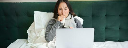 retrato de enfermo asiático mujer acecho videos en computadora portátil, quedarse en cama y tos, atrapando frío foto