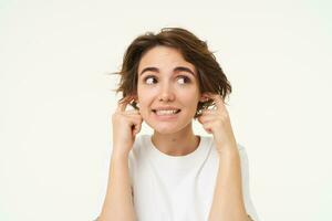 Close up portrait of woman smiling with awkward face expression, shuts her ears, doesnt want to listen, disturbed by loud noise, stands over white studio background photo