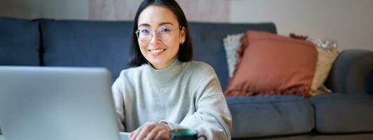 Self-employed young smiling woman, freelancer staying at home, working on remote from laptop, wearing glasses, sitting in living room photo