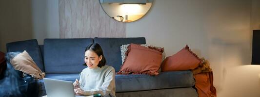 Vertical shot of asian girl sits on floor at home, working on laptop, studying at cozy place, using computer to freelance photo