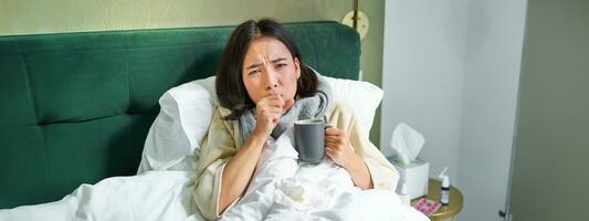 Portrait of couching asian girl in bed, feeling sick, catching cold and staying at home, looking unhealthy, drinking hot tea photo