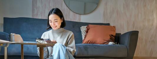 Technology and people. Young stylish asian woman sits at home with her smartphone, texting message, using application on mobile phone photo