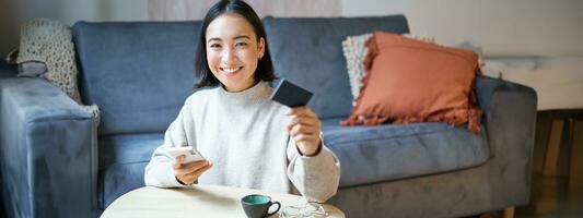 sonriente linda asiático mujer utilizando crédito tarjeta y teléfono inteligente, pago cuentas en línea, participación móvil teléfono, mirando a cámara foto