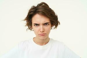 Close up of brunette woman shows angry face, frowning grumpy, pouting and looking disappointed, standing over white background photo