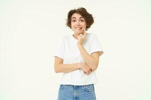 Portrait of cute and shy young woman, looking unsure at camera, posing in white t-shirt against studio background photo