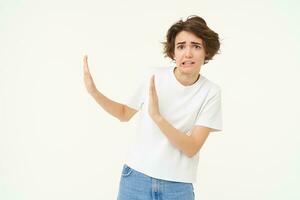 Portrait of awkward, displeased woman shows hand block gesture to the left, avoiding something, refusing, standing over white background photo