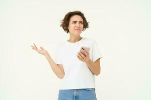 Portrait of woman with smartphone standing confused, puzzled, cant understand something online, using mobile phone, posing over white studio background photo