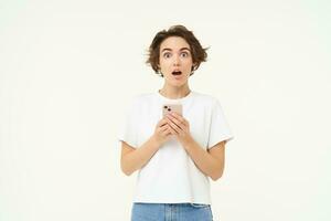 Portrait of brunette girl with shocked face, reading something amazing on mobile phone, standing with smartphone over white background photo