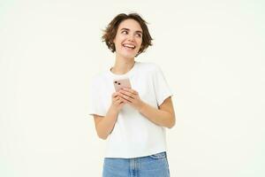 Portrait of young brunette woman texting, sending a message on telephone, using mobile phone and smiling, standing over white studio background photo