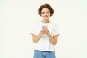 Portrait of young brunette woman texting, sending a message on telephone, using mobile phone and smiling, standing over white studio background photo