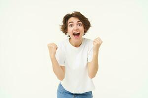 Cheerful brunette woman winning, triumphing, celebrating victory, achieves goal, stands over white background photo