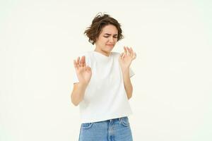 Portrait of woman looking with dislike, rejecting something, raising hands up in defensive gesture, blocking or refusing, standing over white background photo