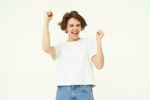 Excited brunette girl, student winning prize, celebrating victory, triumphing, standing in white t-shirt and jeans over white background photo