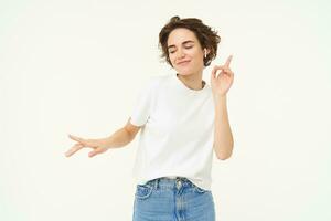 Portrait of carefree, happy young woman in wireless headphones, listens to music, dances to favourite song in earphones, posing over white studio background photo