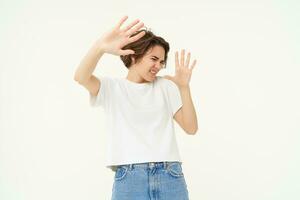 Portrait of woman raising hands in defense and blocking face from something, standing over white background photo