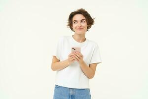 retrato de linda moderno mujer con móvil teléfono, sonriente, utilizando teléfono inteligente aplicación, haciendo en línea compras en solicitud, en pie en contra blanco antecedentes foto