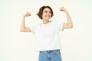 Young happy girl feeling strong, woman shows biceps, flexing muscles on arms and smiling, proves her strength, stands over white background photo