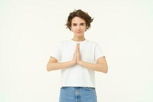 Portrait of mindful young woman, showing namaste, grateful gesture, saying thank you, asking for something and smiling, standing over white background photo