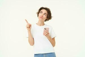 Portrait of young modern woman with mobile phone, looking up and pointing at advertisement, standing against white studio background photo