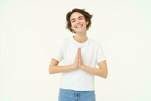 Portrait of brunette young woman holds hands together, namaste gesture, say thank you, grateful, express gratitude and joy, stands over white background photo