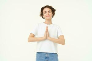 Portrait of mindful young woman, showing namaste, grateful gesture, saying thank you, asking for something and smiling, standing over white background photo