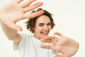 Portrait of happy, laughing young woman, blocks herself from camera, looking joyful, stretching her hands forward, posing over white background photo