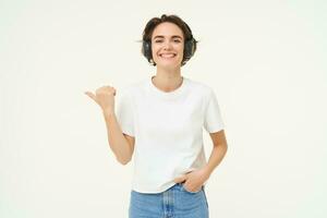 Image of young modern woman, wearing white t-shirt, pointing left, smiling and looking happy, showing advertisement, standing over white background photo