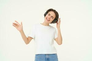 Happy girl dancing and having fun, listens to music in wireless headphones, stands over white background photo