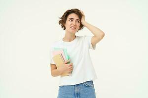 Brunette girl looks troubled, student with problems, holds notebooks and homework with confused face expression, white studio background photo