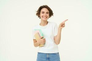 Portrait of brunette woman laughing, student with notebooks pointing at upper right corner, showing banner or advertisement, standing over white studio background photo