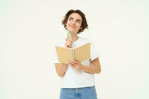 Portrait of creative girl, student doing homework, writing in notebook, standing with her diary, posing over white background photo