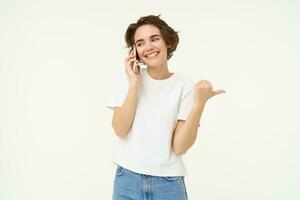 Portrait of young woman talking on mobile phone, using smartphone and pointing finger right, showing direction, banner advertisement, studio background photo