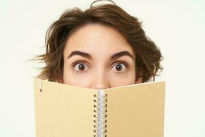 Close up portrait of girl face, hiding behind notebook, looking aside, standing over white background photo
