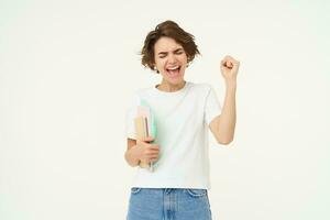 Excited and enthusiastic girl, feeling thrilled, makes fist pump and screams from joy, posing with notebook against white studio background photo