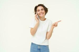 retrato de joven mujer hablando en móvil teléfono, utilizando teléfono inteligente y señalando dedo bien, demostración dirección, bandera anuncio publicitario, estudio antecedentes foto