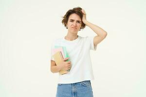 Brunette girl looks troubled, student with problems, holds notebooks and homework with confused face expression, white studio background photo