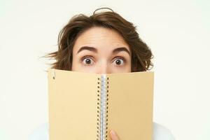 Close up portrait of girl face, hiding behind notebook, looking aside, standing over white background photo