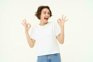 Portrait of young woman, showing okay, ok gestures and smiling, giving positive feedback, recommending, standing over white background photo