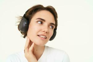 Image of young modern woman in wireless headphones, using earphones to listen to music, standing over white background photo