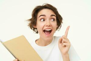 Portrait of happy young woman with planner, student with homework notes, pointing at upper right corner, showing banner and making amazed face expression, standing over white background photo