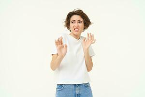 Young reluctant woman steps back, raises hands to refuse, rejects something disgusting, reaction of aversion and dislike, isolated over white background photo