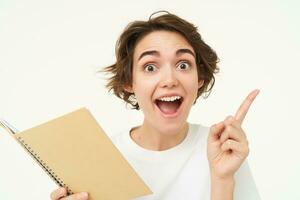 Enthusiastic brunette woman with diary, holding planner book, pointing at upper right corner and smiling with excitement, standing over white background photo