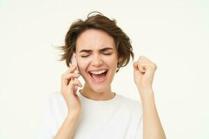 Excited brunette woman celebrating, answers phone call and looking happy, winning, triumphing from great big news, standing over white background photo