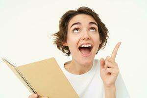 Enthusiastic brunette woman with diary, holding planner book, pointing at upper right corner and smiling with excitement, standing over white background photo