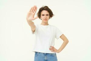 Image of serious young woman extends one hand, shows stop, prohibit gesture, forbid something, standing over white background photo