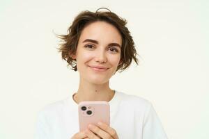 cerca arriba retrato de joven mujer sonriente, utilizando teléfono inteligente, enviar mensajes de texto, comprobación su móvil aplicaciones, soportes terminado blanco antecedentes foto