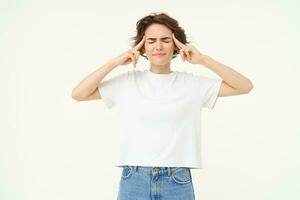 Image of brunette woman touches her head and frowns, looks anxious, thinking, trying to remember, looking worried and concerned, standing over white background photo